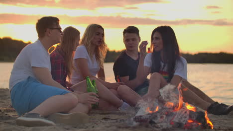 Five-students-are-sitting-in-shorts-and-T-shirts-around-bonfire-on-the-sand-beach.-They-are-talking-to-each-other-and-drinking-beer-at-sunset.